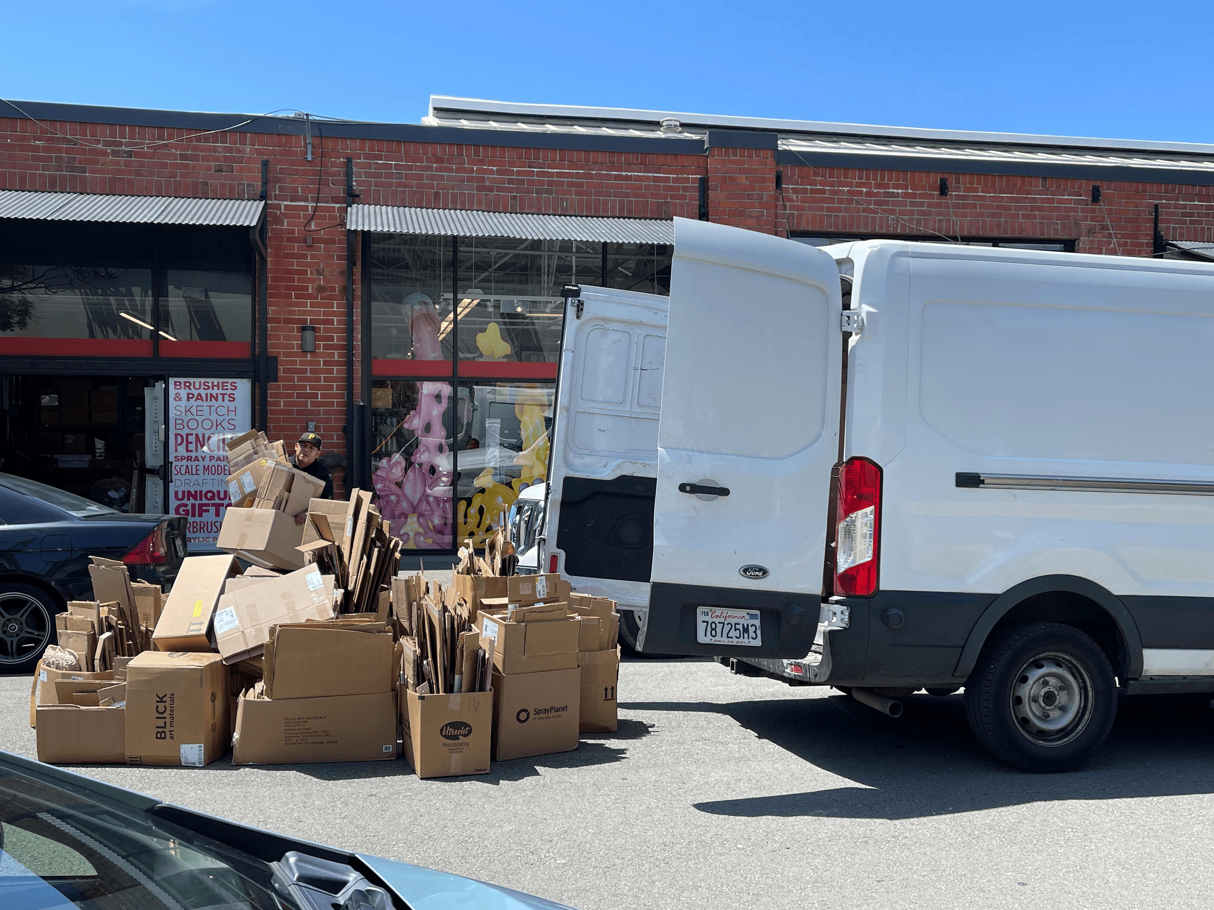 A white van with its rear doors open is parked on a street, surrounded by a large pile of flattened cardboard boxes.