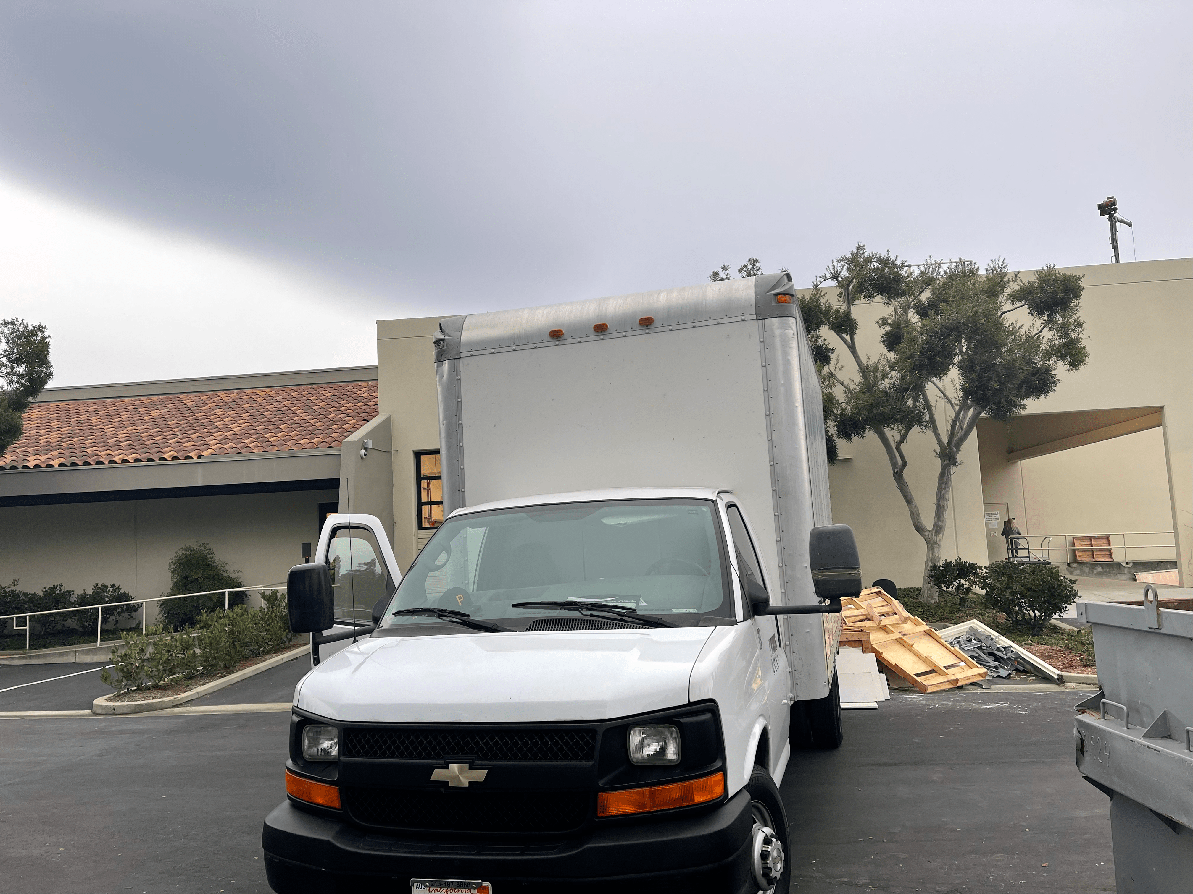 A white Chevrolet Express box truck is parked in a parking lot with buildings and overcast sky in the background.